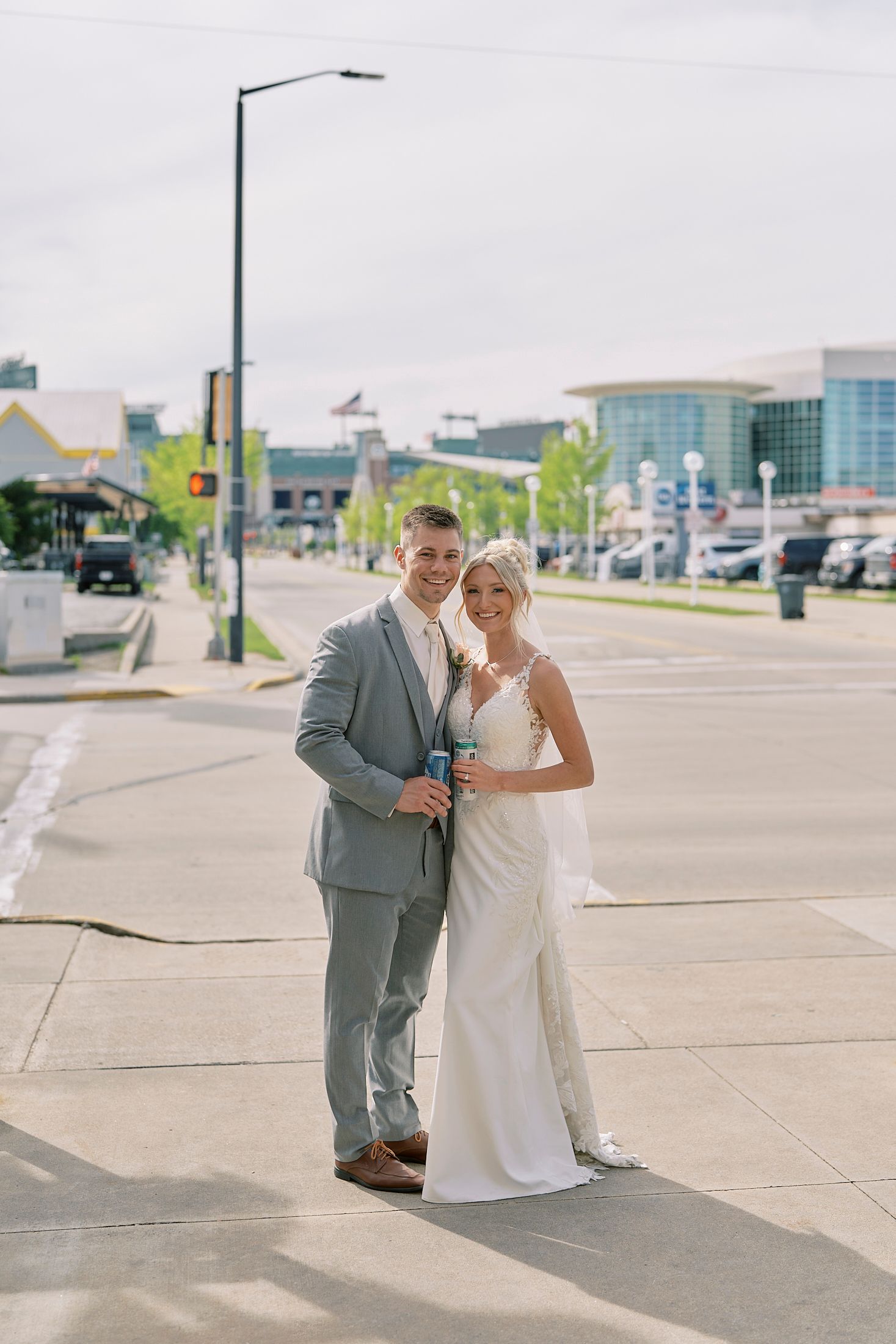 Bride and groom posing on a city street, she in a white gown, he in a gray suit, holding drinks.