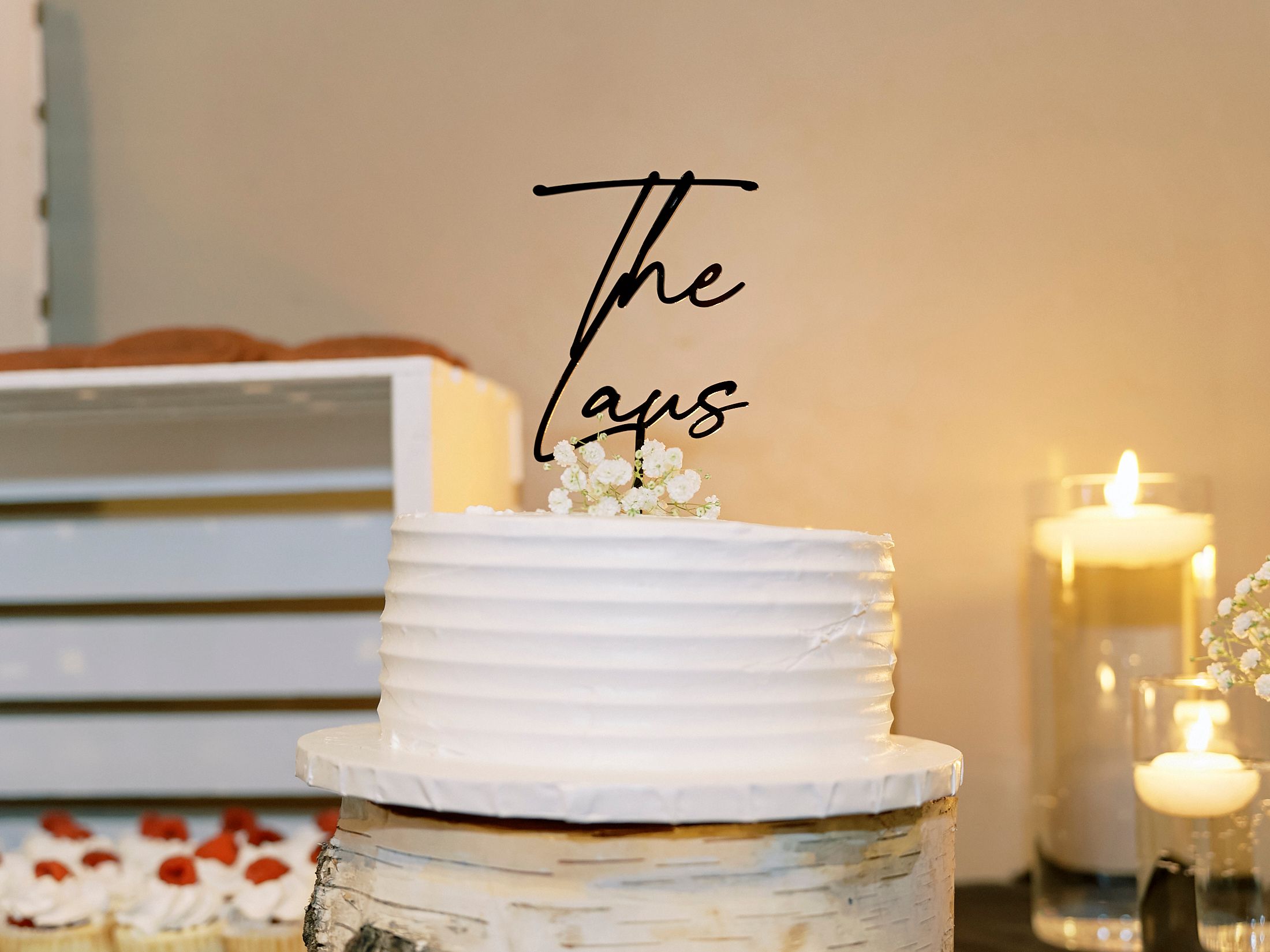 Elegant white wedding cake topped with a black acrylic 'The Taus' cake topper, surrounded by candles.