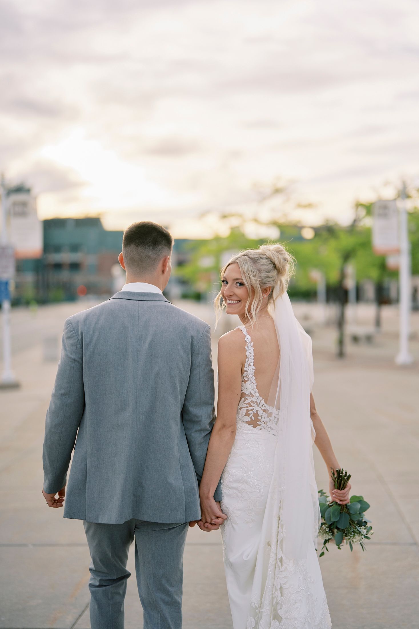 Bride in lace back wedding dress holding bouquet, smiling back at camera with groom at sunset.