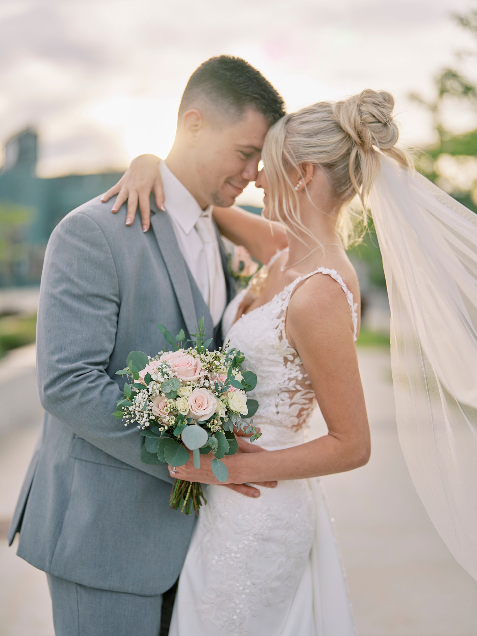 Bride and groom sharing romantic forehead touch at sunset, bride holding blush rose bouquet.