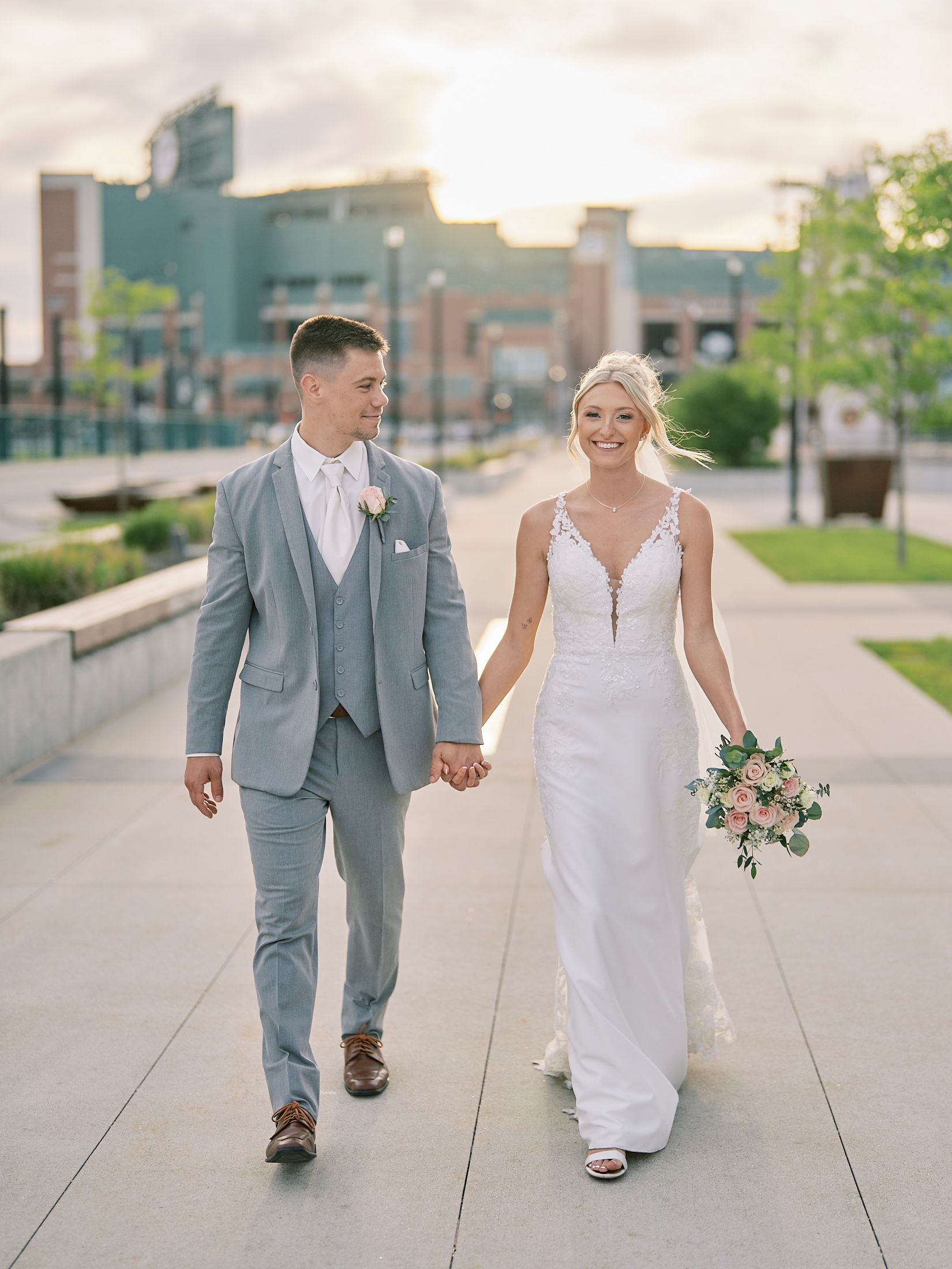 Smiling bride and groom walk hand-in-hand at golden hour, bride holding a pink bouquet in a fitted gown.
