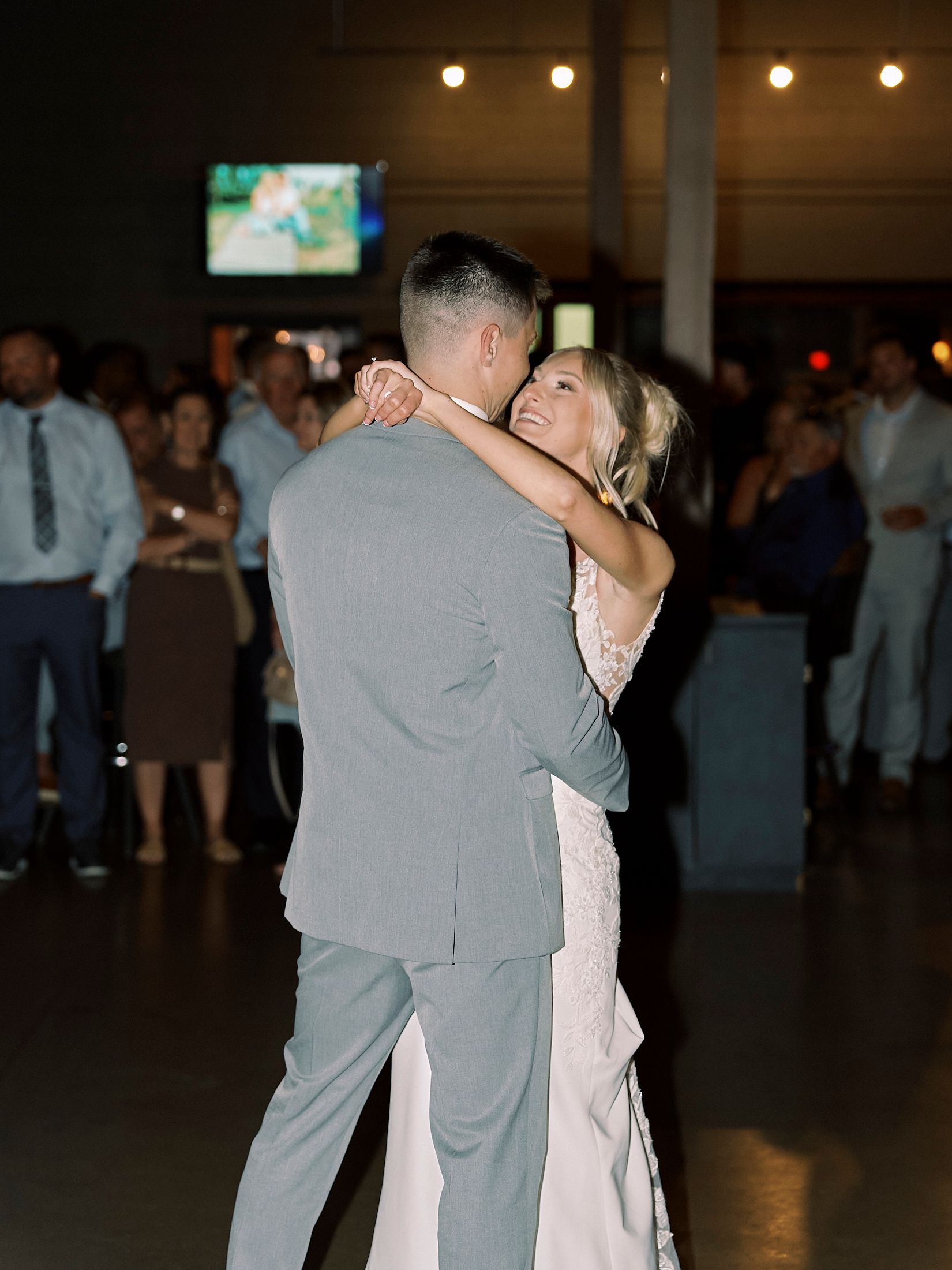 Bride and groom share their first dance, surrounded by guests at a warmly lit wedding reception venue.