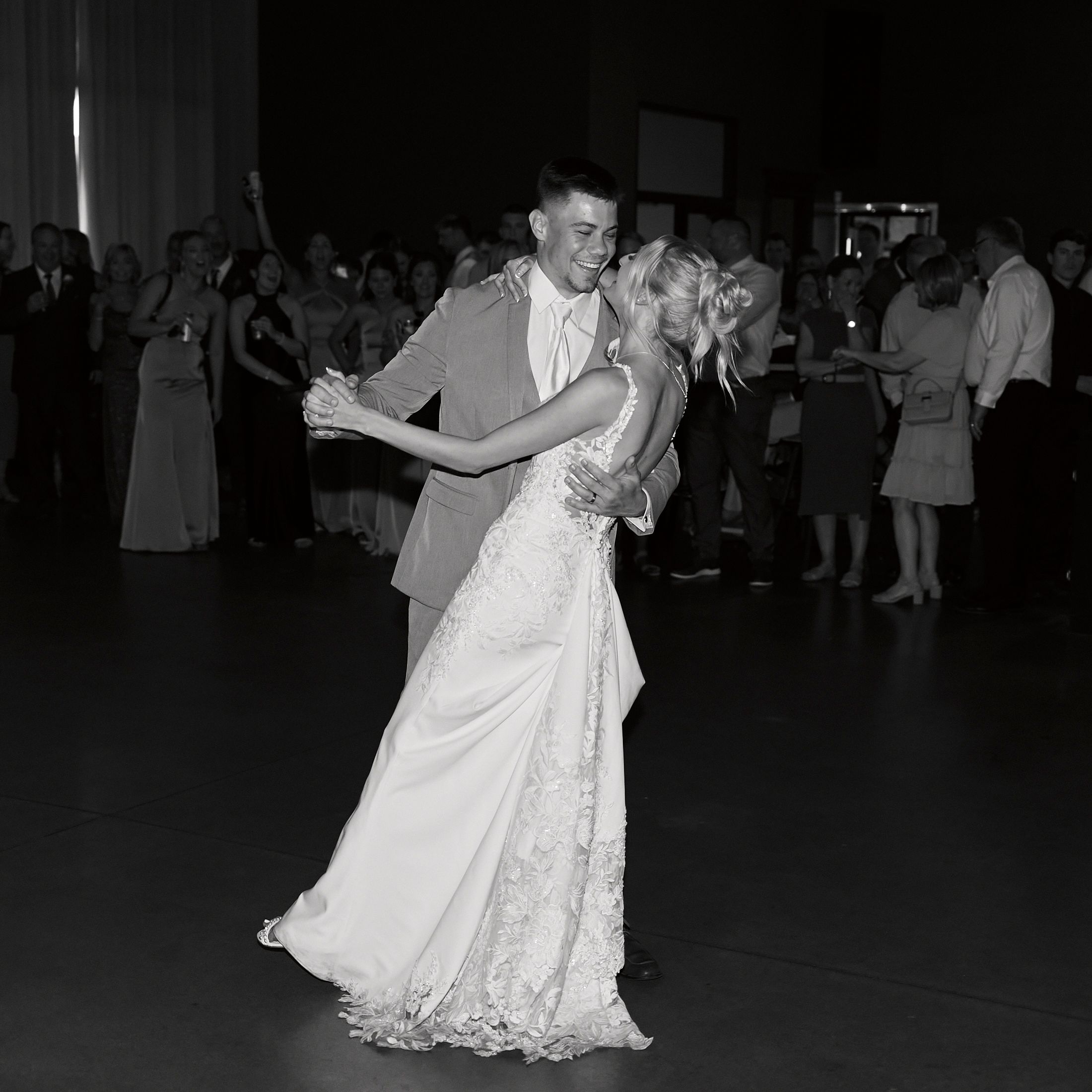 Bride and groom share their first dance surrounded by wedding guests in black and white photo.