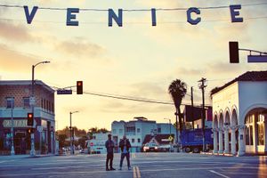 Two people cross a street under the iconic Venice sign at sunset in Venice Beach, California.