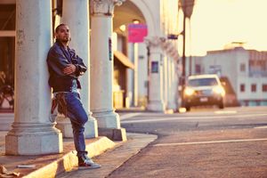 A stylish young man leans against a white column on a sunny urban sidewalk with arms crossed.