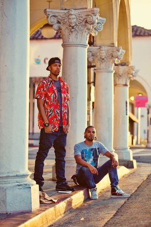 Two young men pose by ornate decorative columns on a sunny sidewalk, one standing and one seated on the curb.