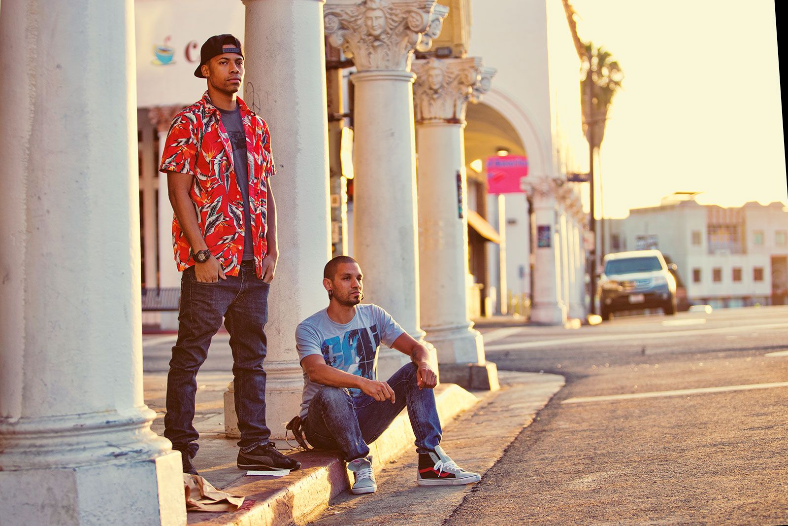 Two young men pose by white ornate columns on a sunny sidewalk, one standing and one sitting on the curb.