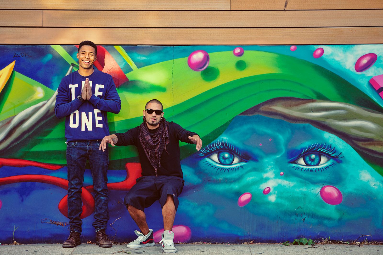 Two young men pose confidently in front of a vibrant colorful graffiti mural featuring a large face.