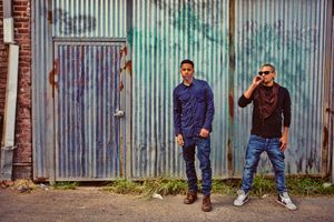 Two men stand in front of a rustic corrugated metal wall in an urban alley setting.
