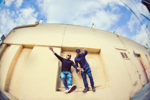 Two men pose dramatically on a rooftop ledge against a bright blue cloudy sky using a fisheye lens.