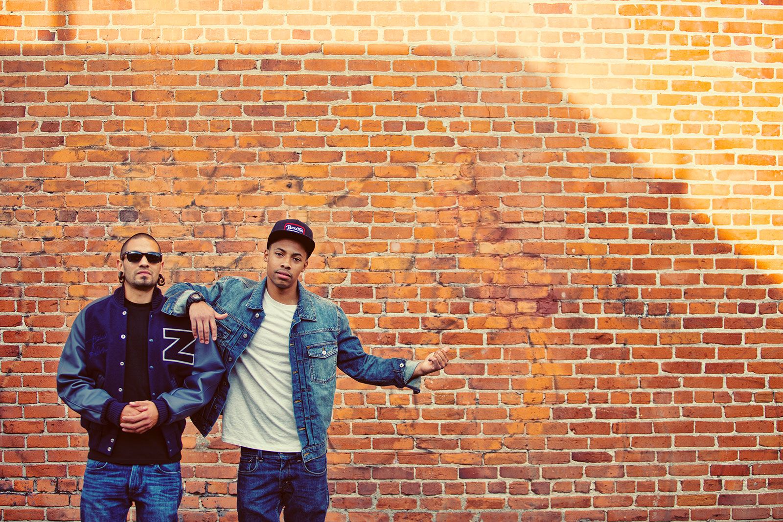 Two stylish men posing against a rustic red brick wall in urban streetwear fashion.