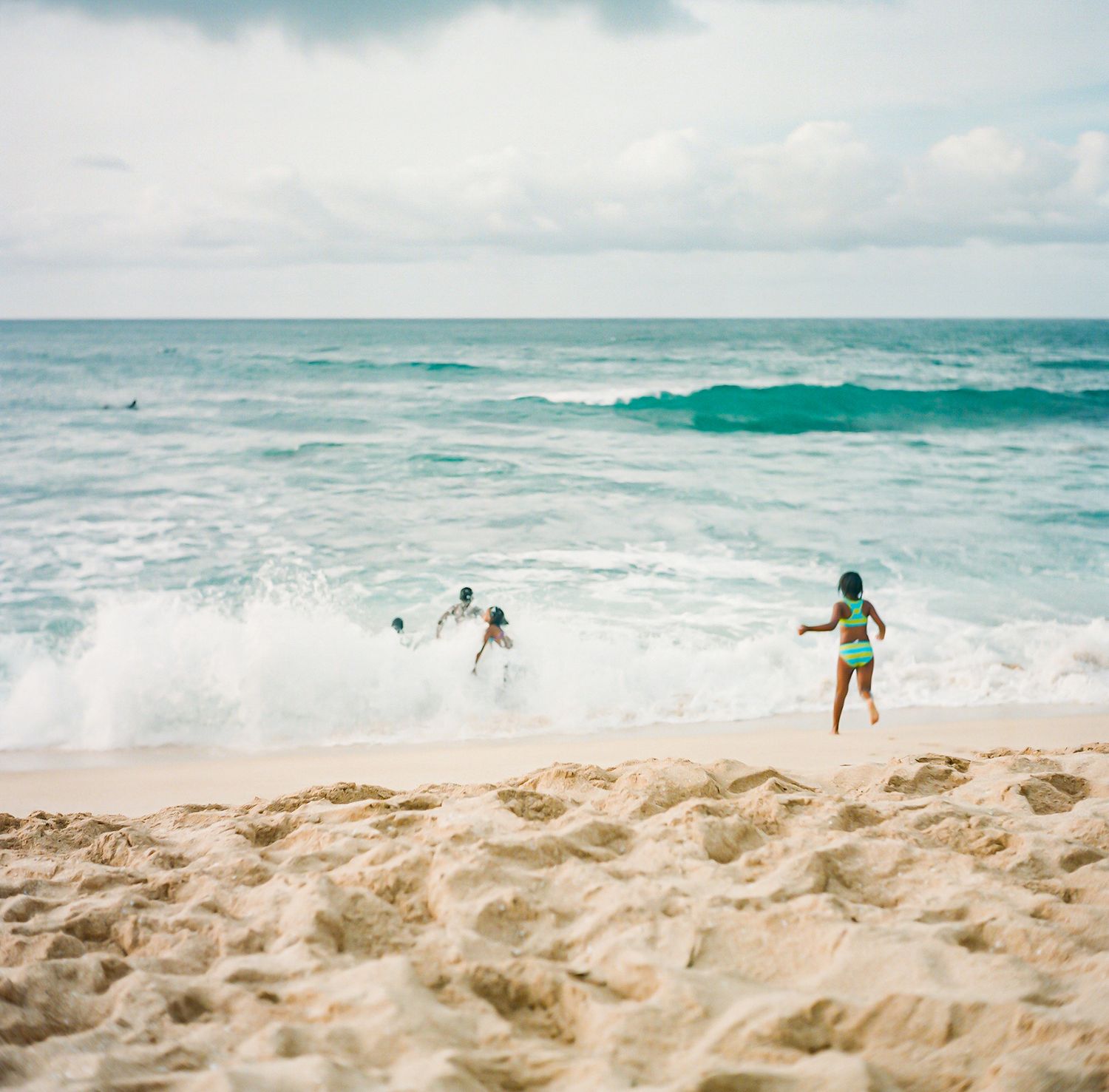 Children running toward turquoise ocean waves on a sandy beach under a partly cloudy sky.