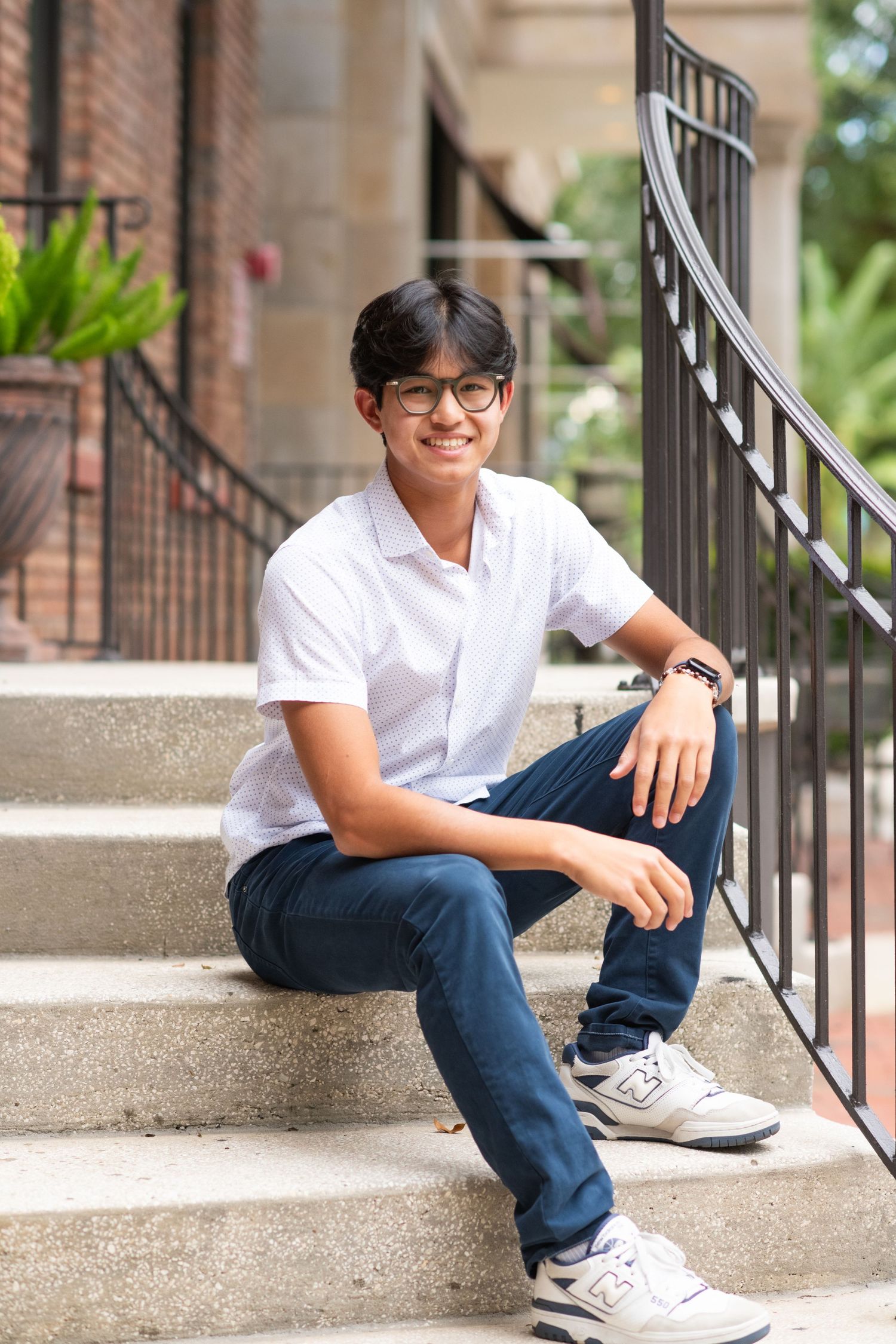 Young man in white polo and navy pants smiling on outdoor steps, wearing glasses and sneakers.