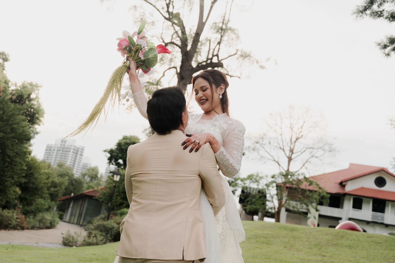 A joyful bride in a white lace gown is lifted by her groom while holding a pink bouquet outdoors.
