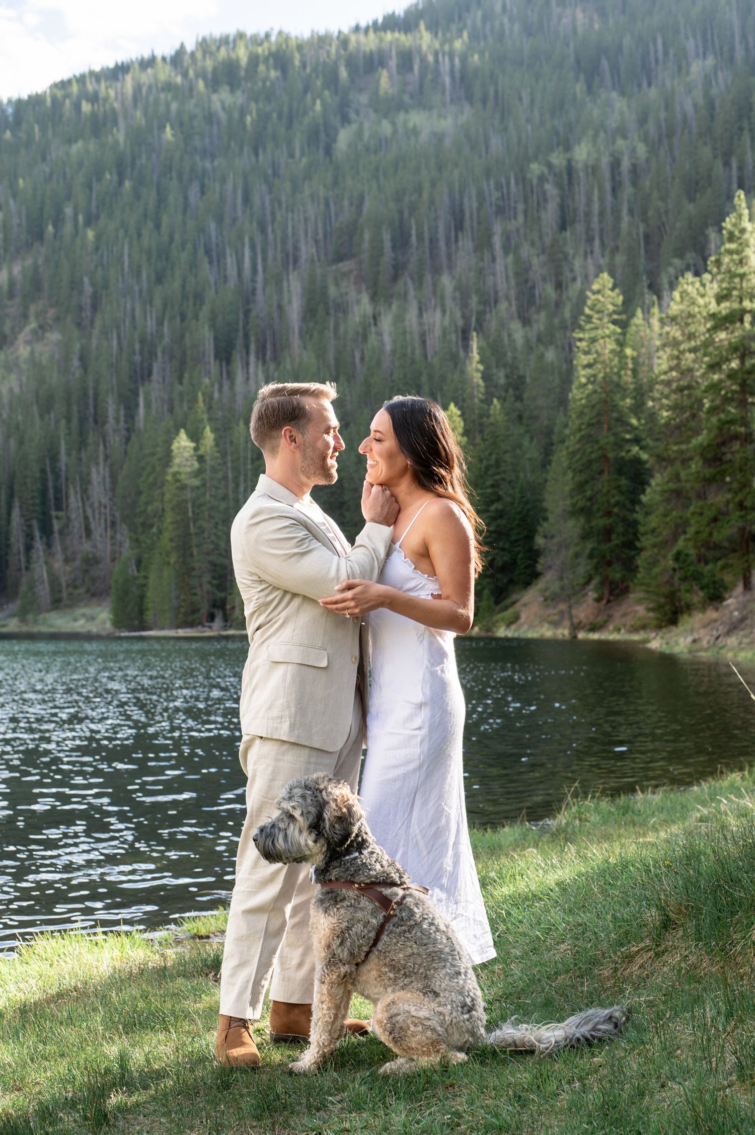 Couple in wedding attire poses by a mountain lake with their fluffy dog, surrounded by pine trees for an elopement at Officer's Gulch