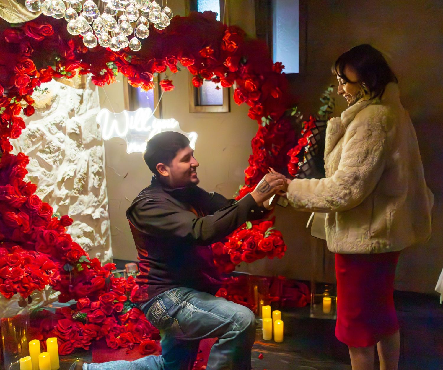 Man proposes to woman on one knee surrounded by red roses heart arch and candles in romantic setting.