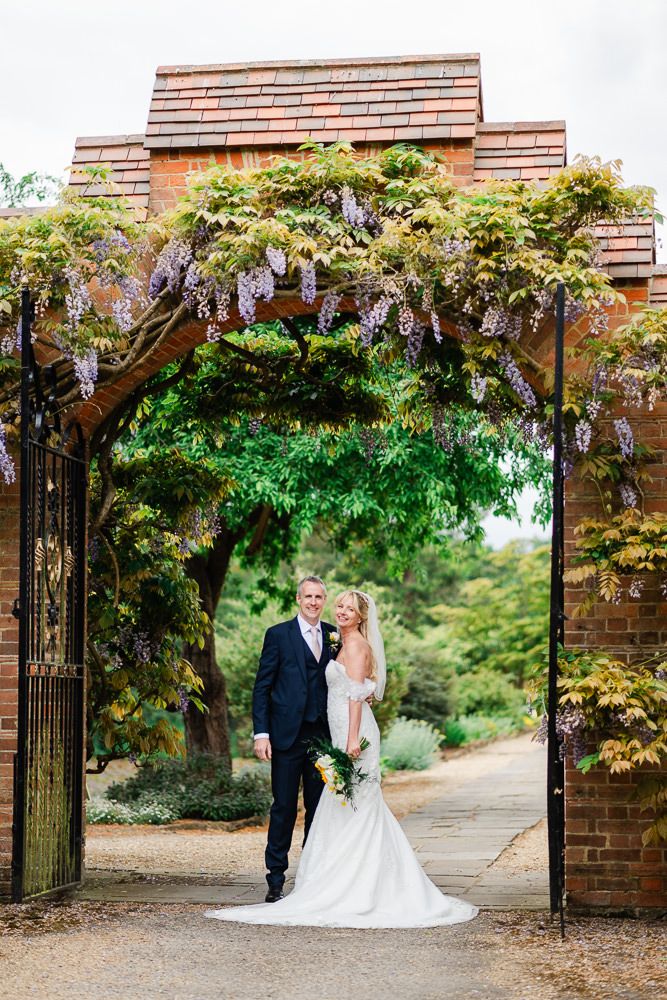 Bride and groom pose under a wisteria-draped brick archway with iron gates in a lush garden setting.