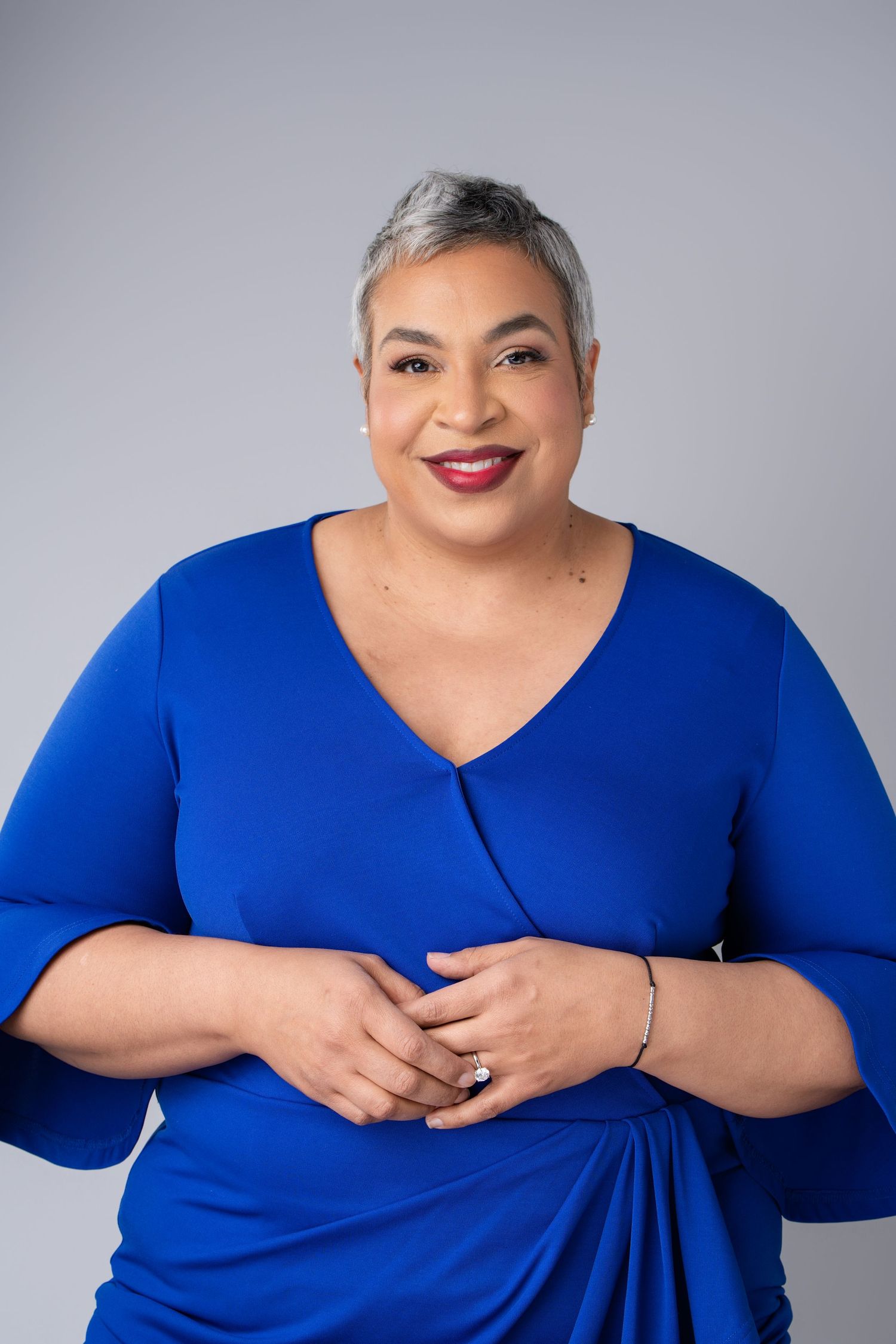 Woman in blue wrap dress with short gray hair smiling confidently against gray background.