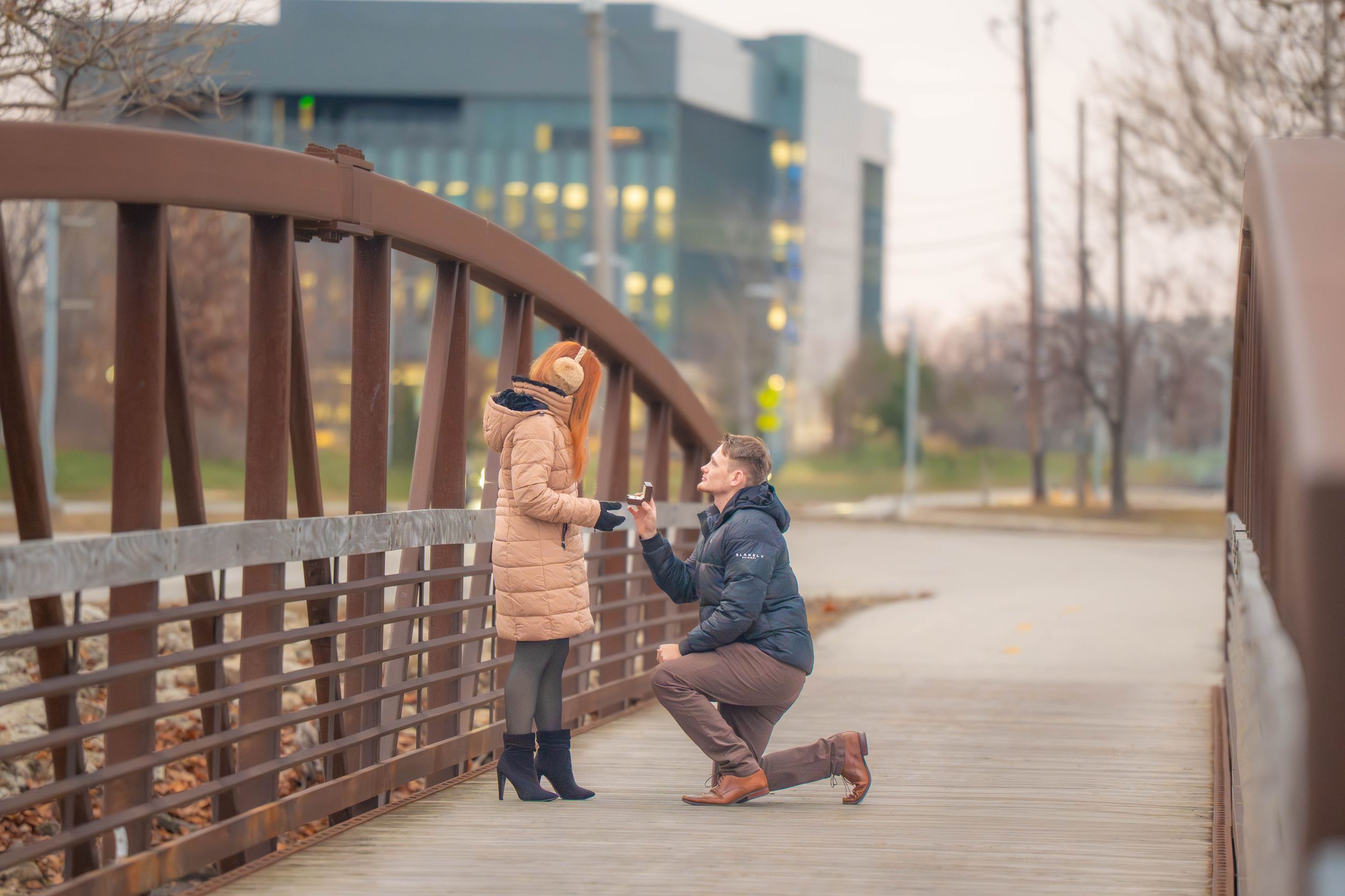 Man proposes to woman on a bridge, kneeling with ring box while she covers her mouth in surprise outdoors. Quad Cities Mississippi River