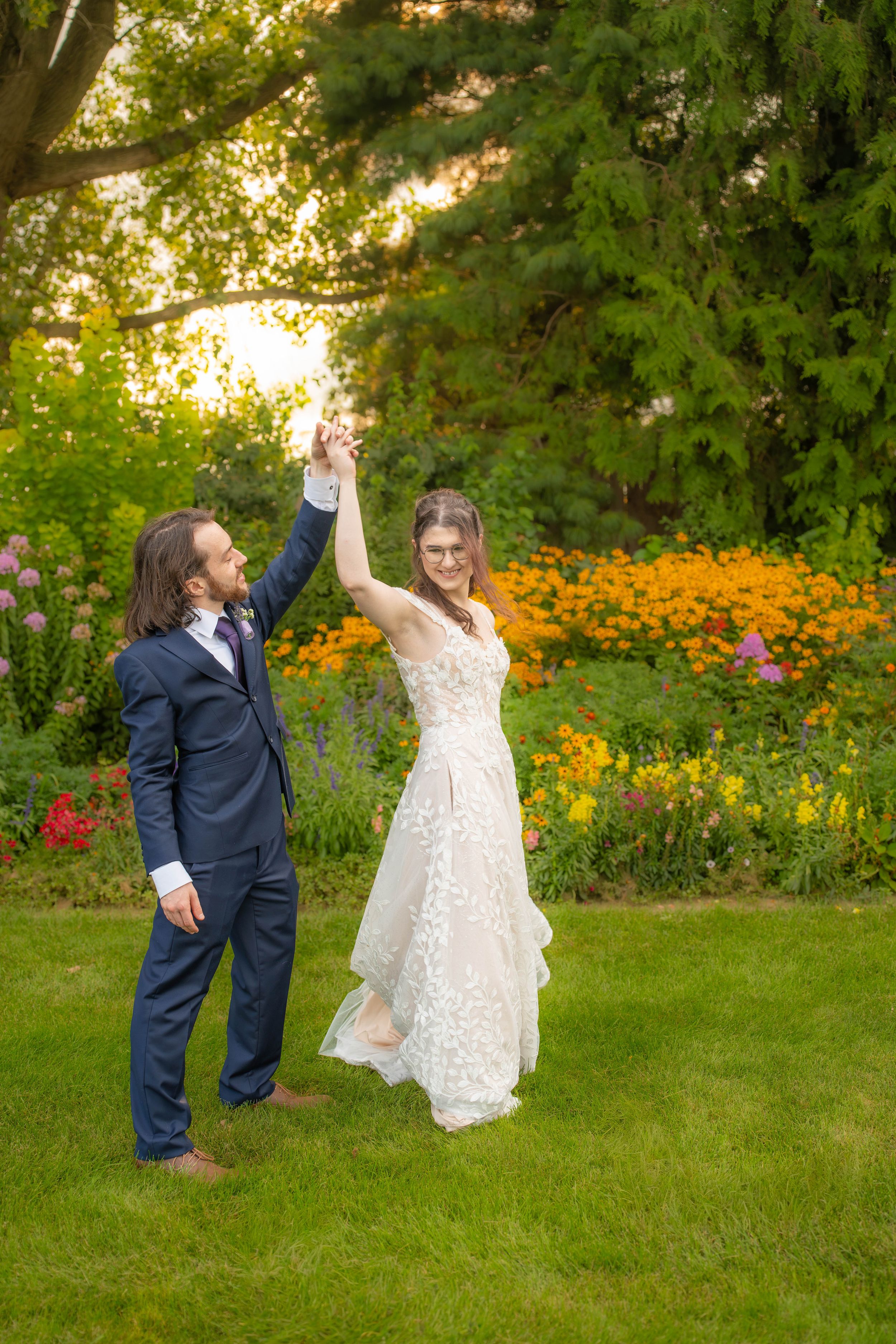 Bride and groom dancing outdoors in a lush garden, she in lace gown, he in navy suit, hands raised joyfully. Quad Cities Wedding Photography