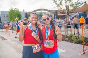 Two women wearing 5k Firecracker Run race medals and bibs smile at a finish line event on a sunny day. Quad Cities Event Photography