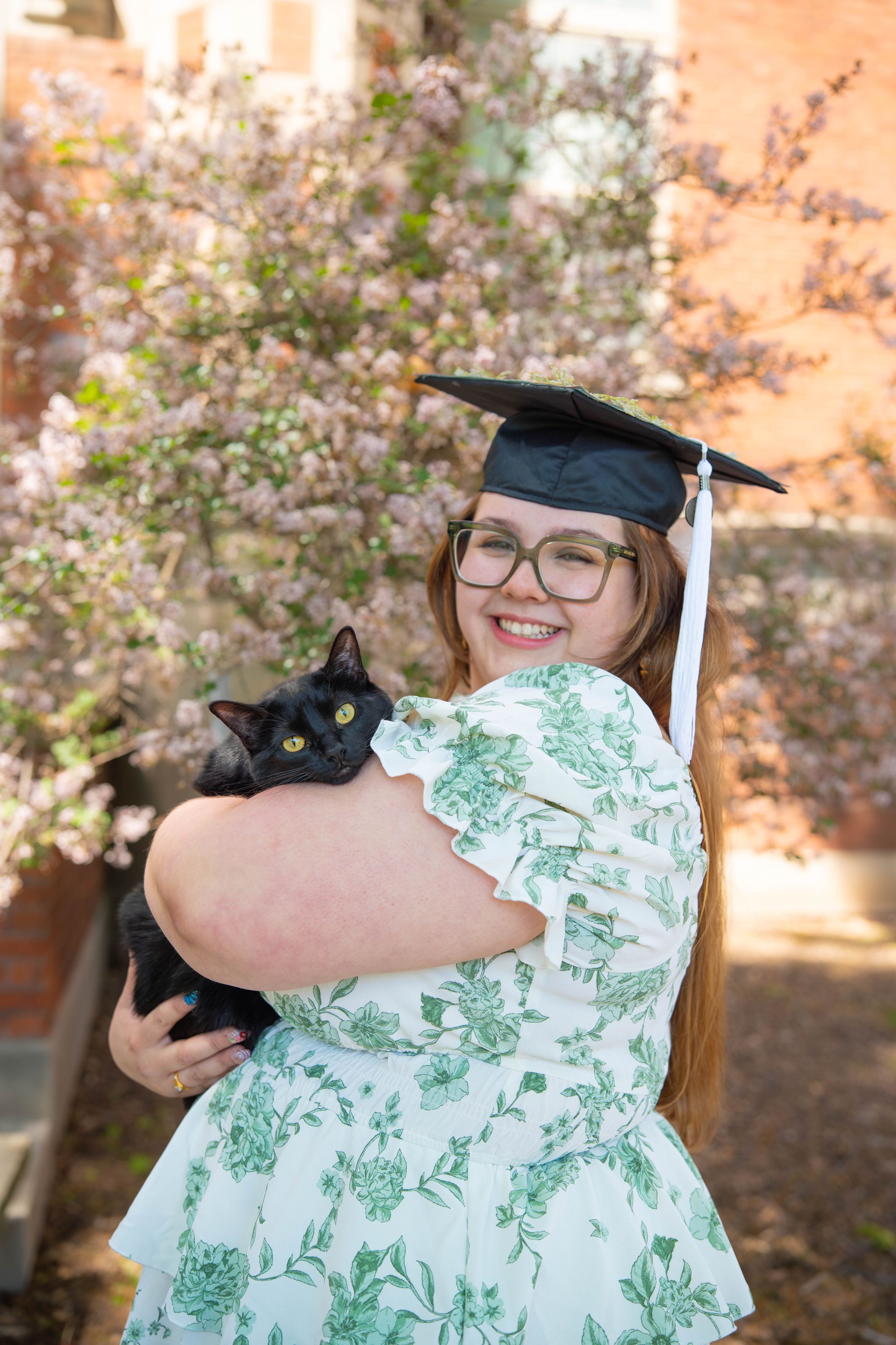 Smiling graduate in cap and floral dress holds black cat outdoors near blooming trees in spring. Quad Cities Senior Portraits