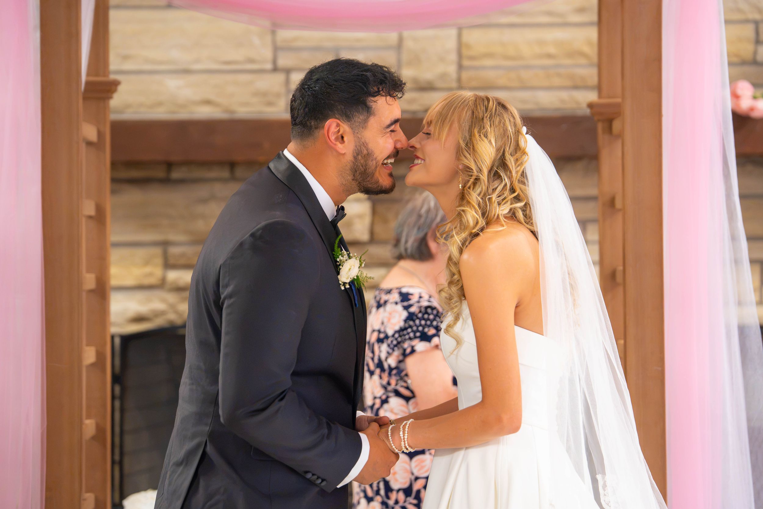 Bride and groom share kiss at wedding ceremony with pink decor and stone fireplace backdrop. Quad Cities Wedding Photography