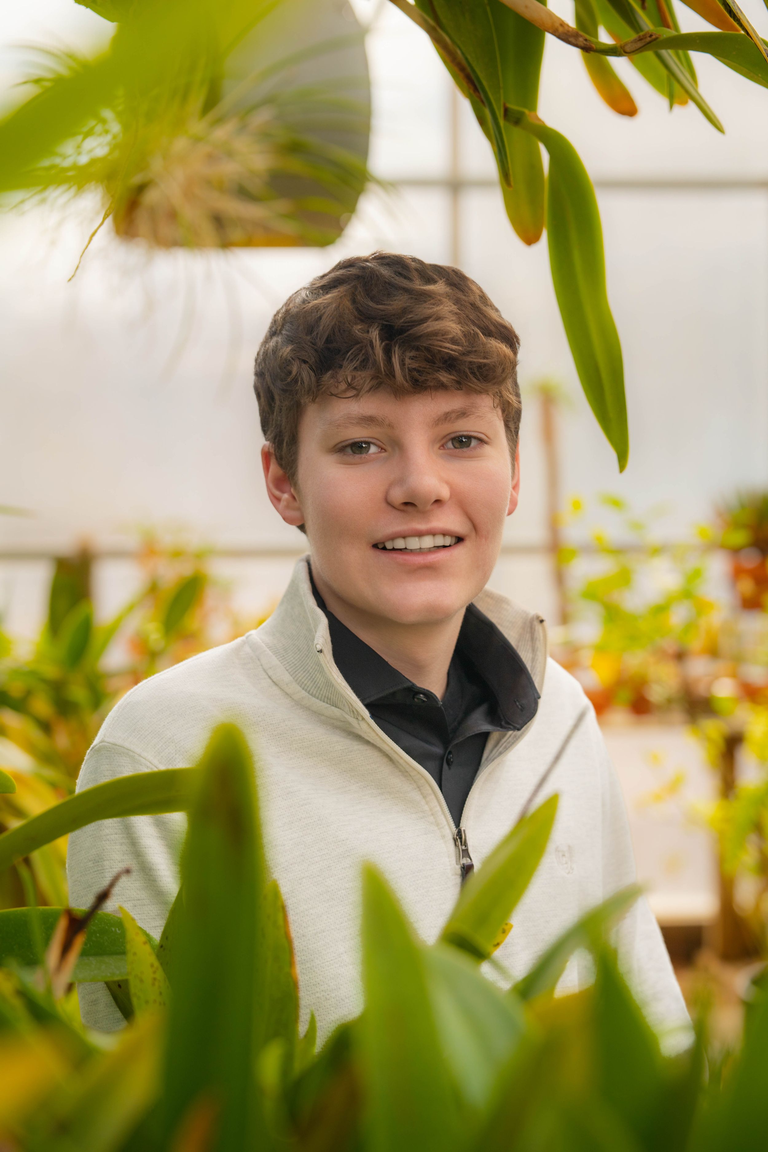 Young person smiling among lush green plants in a bright greenhouse, wearing a white zip-up jacket. Quad Cities Senior Portraits QCBC