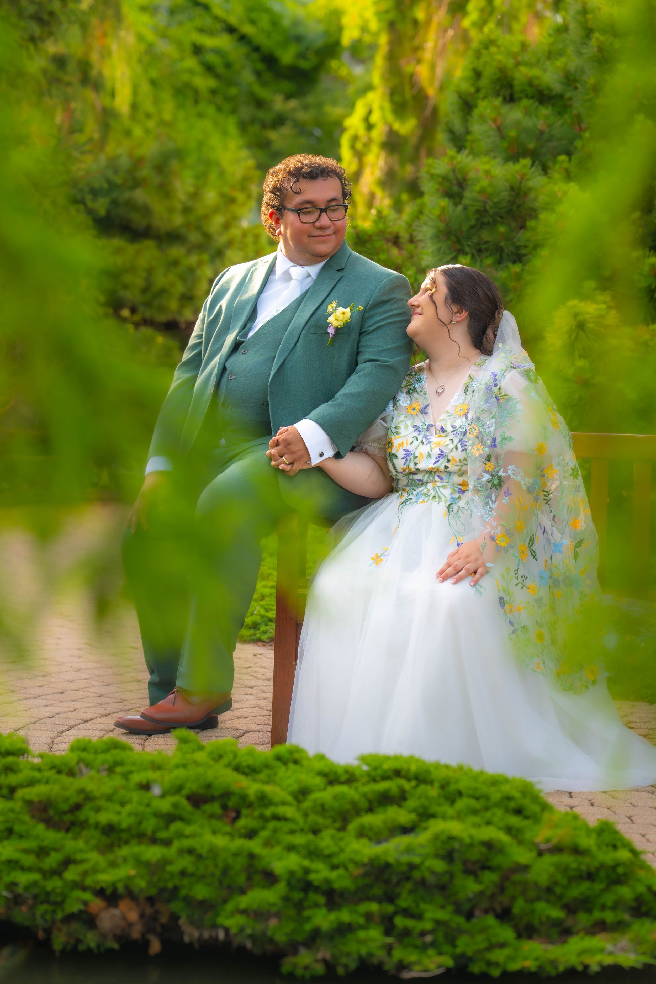 A couple in wedding attire poses in a lush green garden, groom in teal suit, bride in floral white gown. Quad City Botanical Center