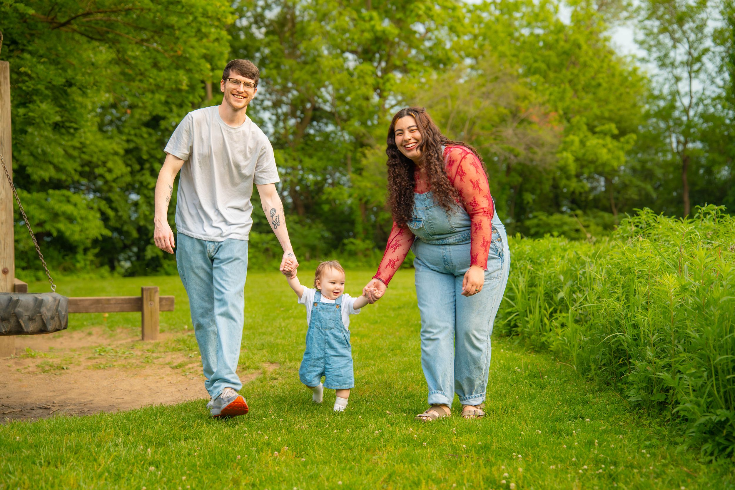 Young family with toddler walking hand-in-hand on green grass path surrounded by lush trees on a sunny day. Quad Cities Blackhawk State Park