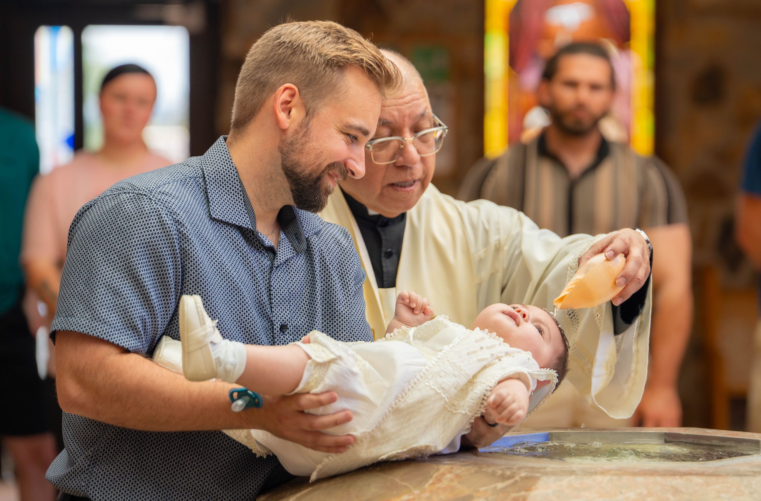 A priest performs a baby baptism ceremony, pouring holy water while a smiling father holds the infant in white. Quad Cities Photography
