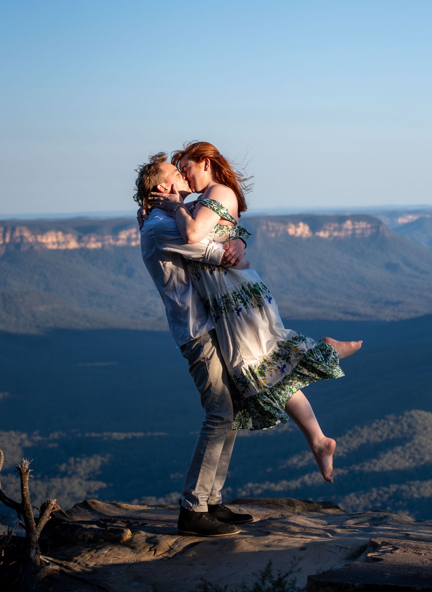 Couple sharing a romantic kiss on a mountain overlook, man lifting woman in floral dress with blue valley views.
