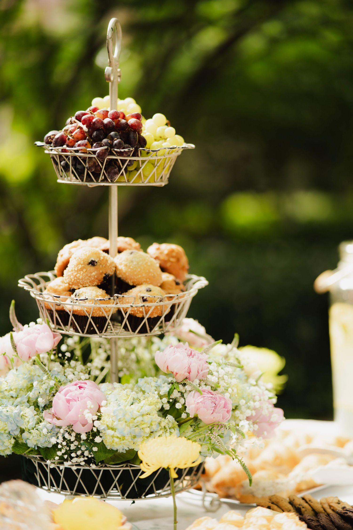 Close-up of elegant three-tiered wire stand with grapes, pastries, and fresh flowers at garden party.