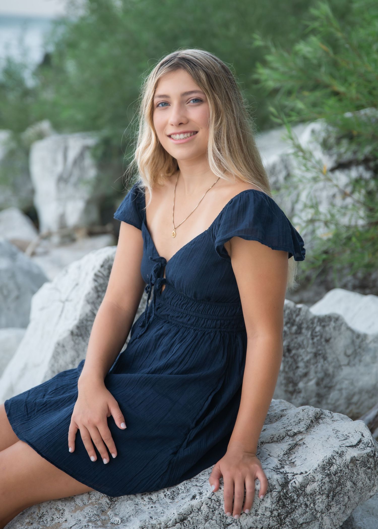 Young woman in navy blue dress sitting on rocks outdoors, smiling, with greenery in background.