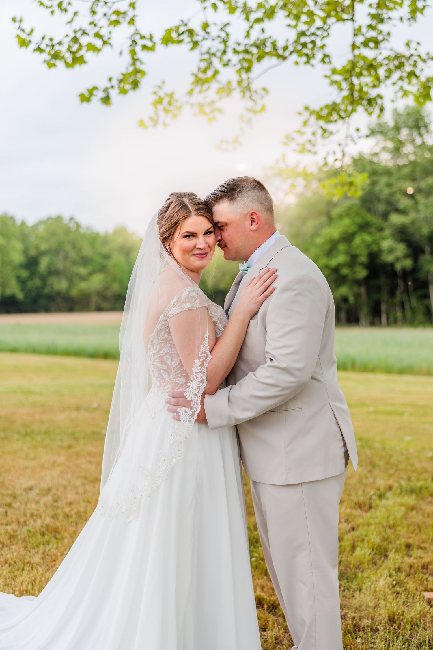 A romantic moment between newlyweds in a sun-lit meadow with green trees in the background.