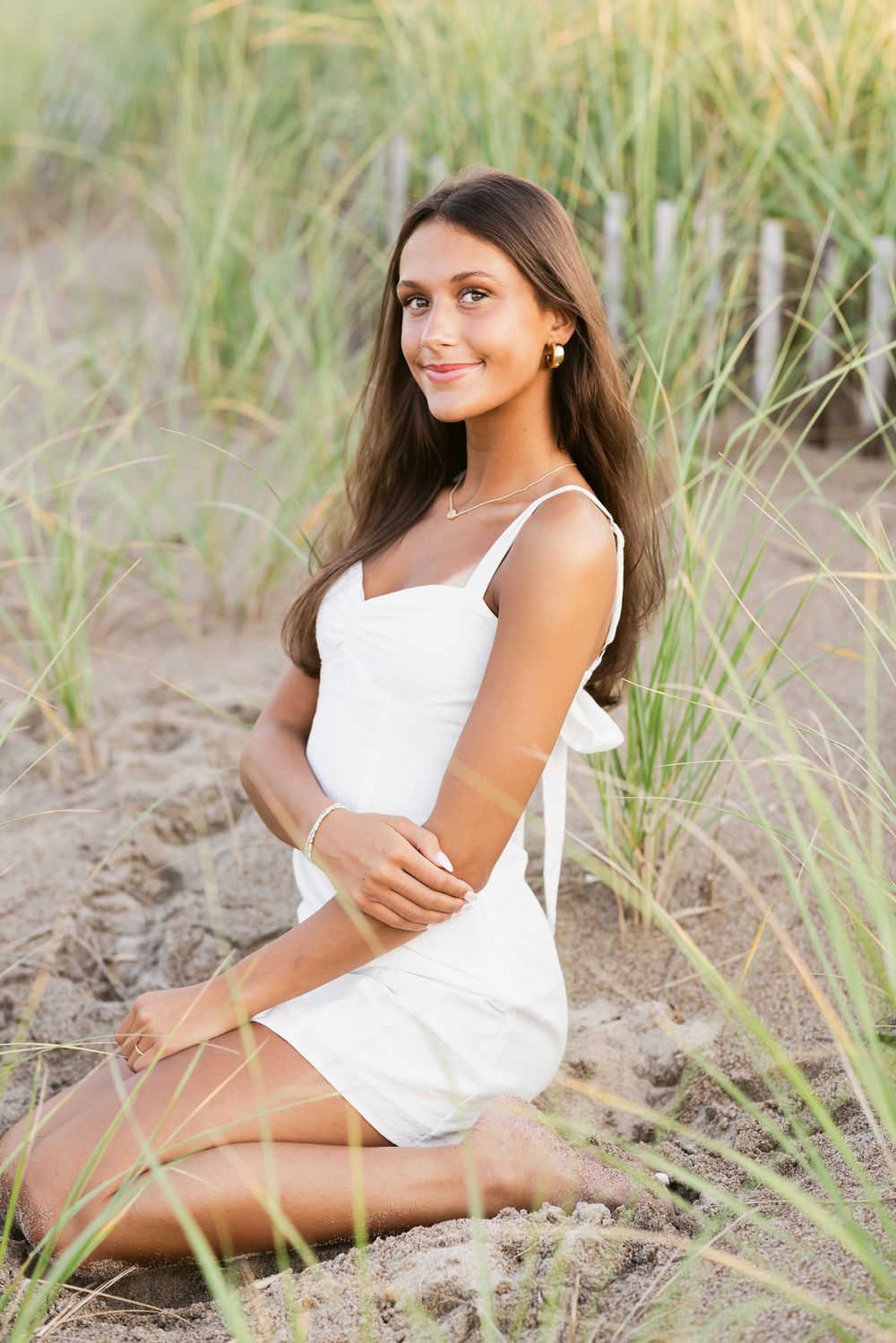 Dark hair girl wearing a short white dress sits in sand in seagrass for senior portraits. Manchester by the Sea senior photographer