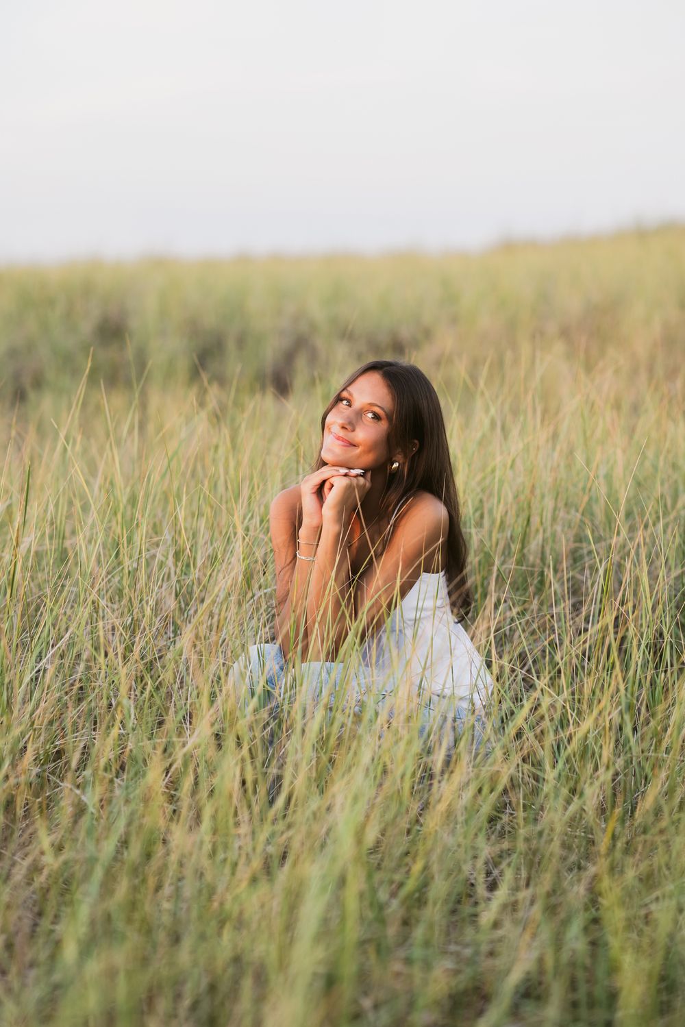 Dark hair teen girl crouches with hands under chin in seagrass for senior portraits wearing a white tank top. Chelmsford senior photographer