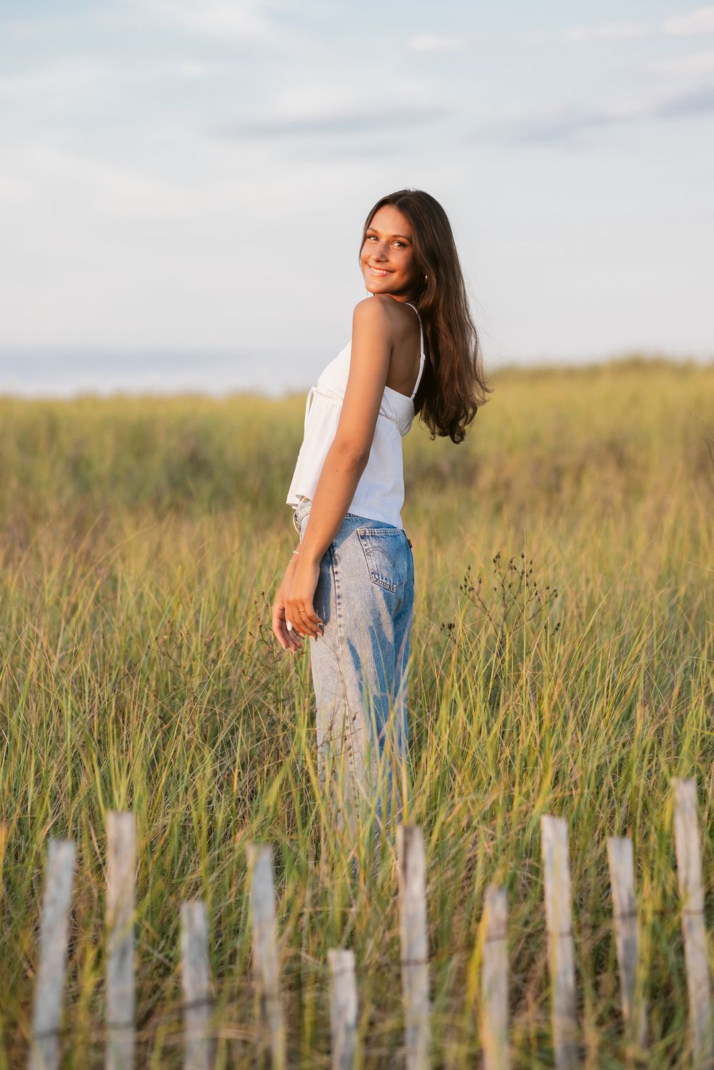 Teen girl wears white tank top and blue jeans poses in coastal grass under blue sunset skies. Manchester senior photographer.