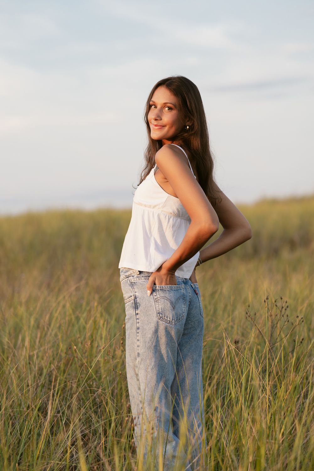 Dark haired teen girl wearing white tank and blue jeans poses with hands in back pockets in coastal grass. Salisbury senior photographer