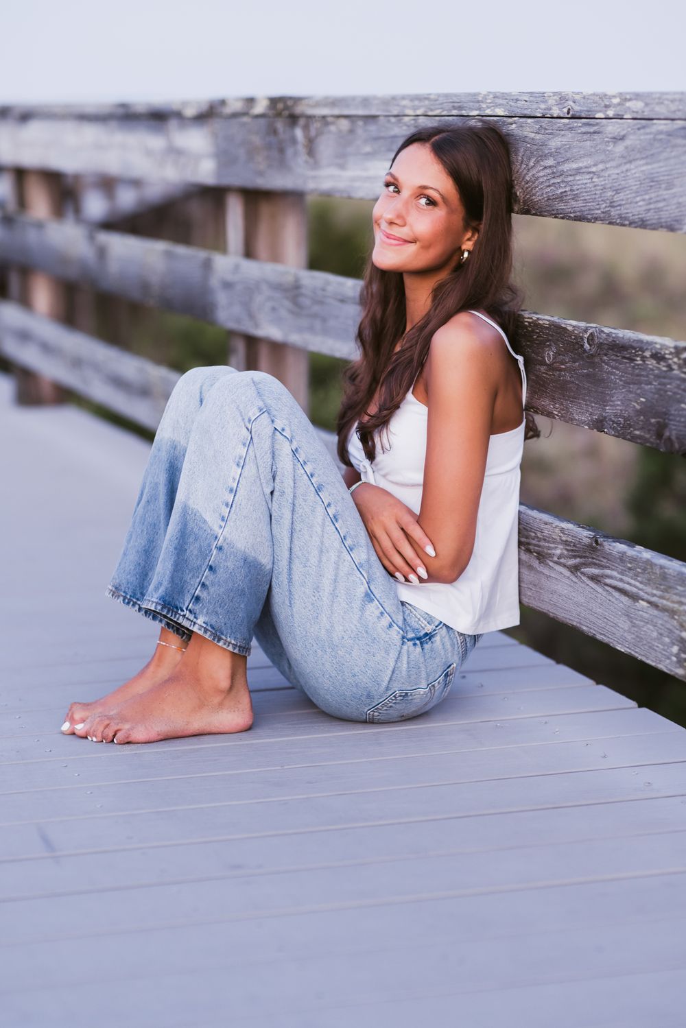 Teen girl with long brown hair sits with knees to chest wearing white tank and blue jeans leaning against a weathered wooden boardwalk