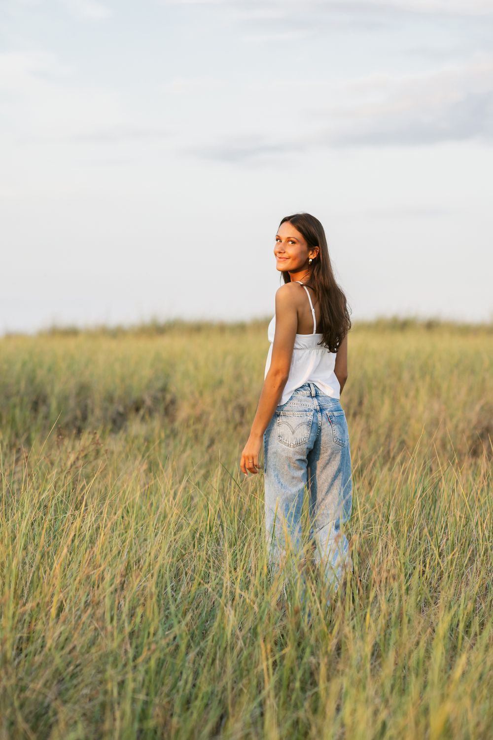 Young lady with long brown hair wearing white tank and blue jeans walks in coastal grass with blue skies above. Andover senior photographer