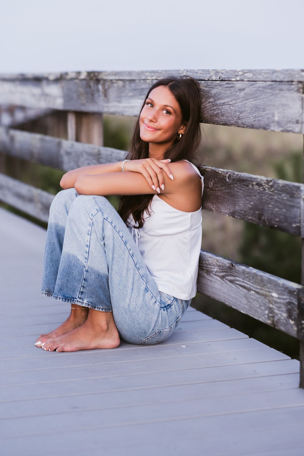 Senior girl with brown hair rests her elbows on knees posing for senior photos on boardwalk at Salisbury Beach. Harvard senior photographer