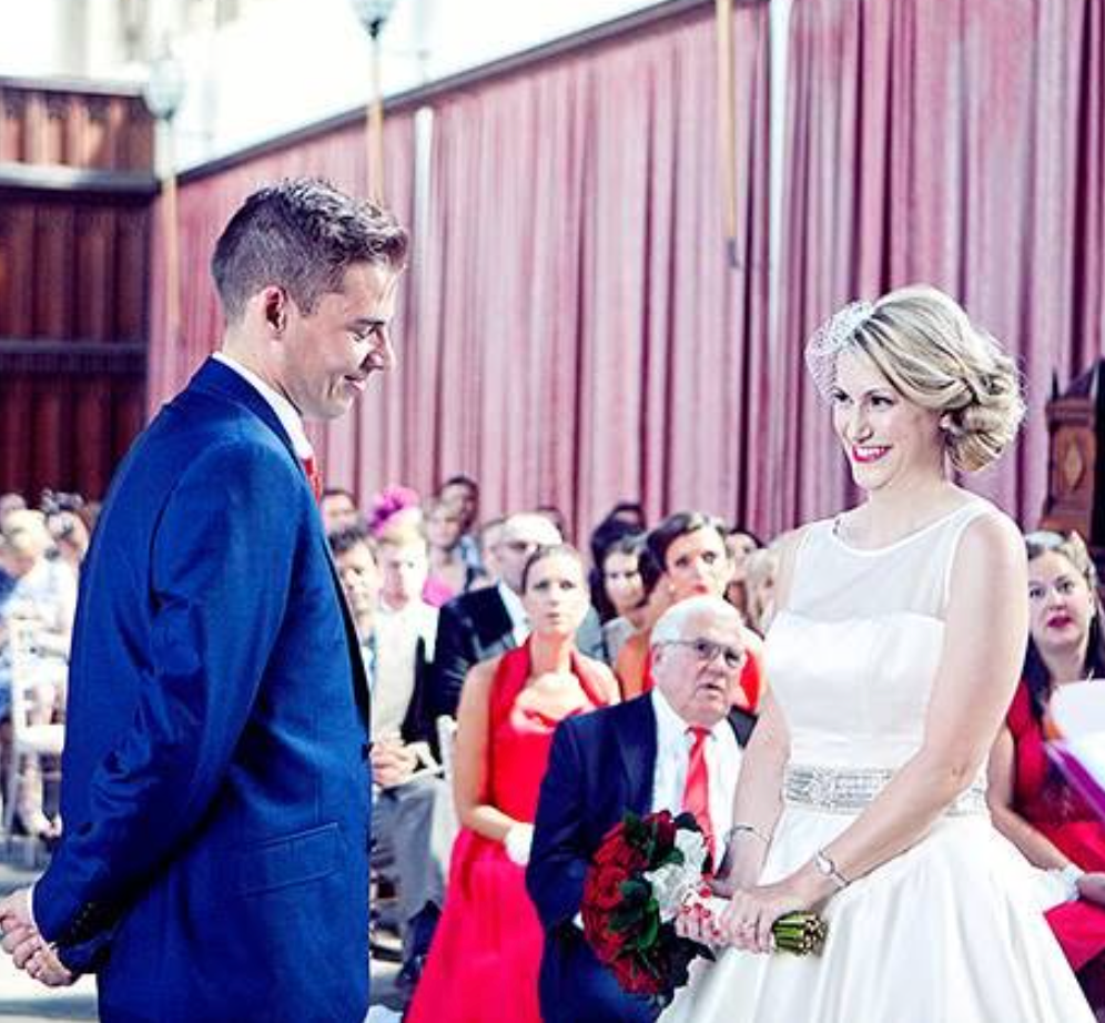 A smiling bride in a white gown and groom in a blue suit exchange vows at an outdoor wedding ceremony.