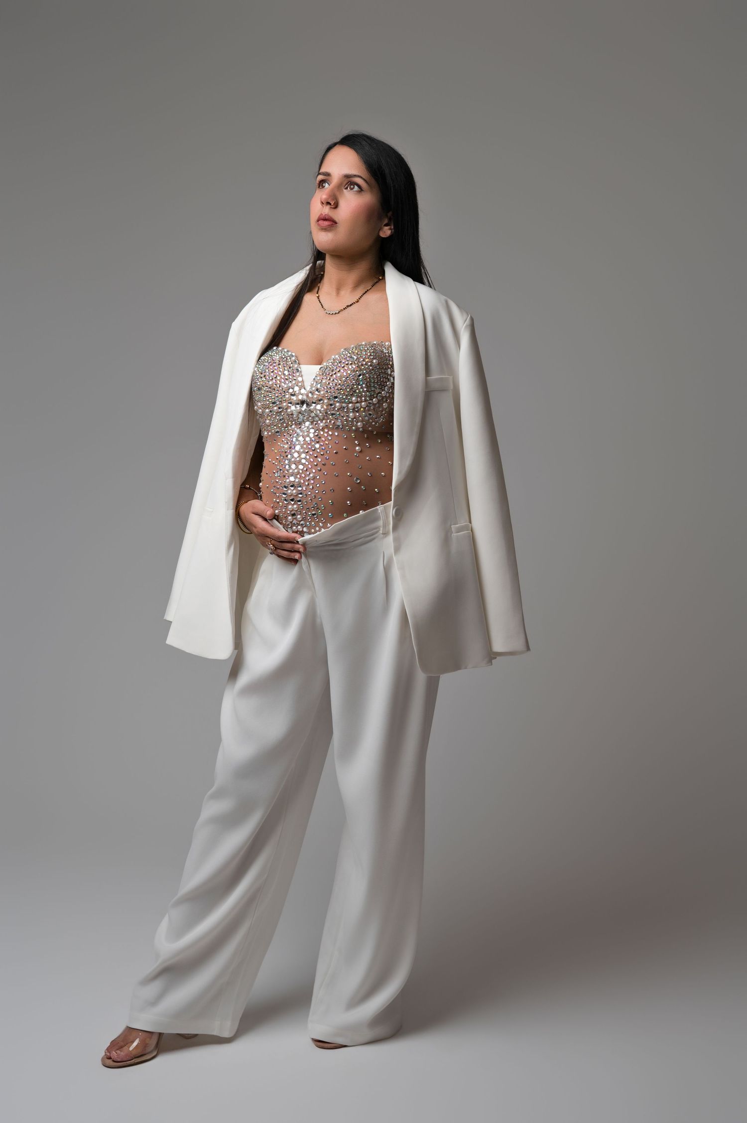 Pregnant woman in white blazer and trousers with silver sequin top, posing on gray background.