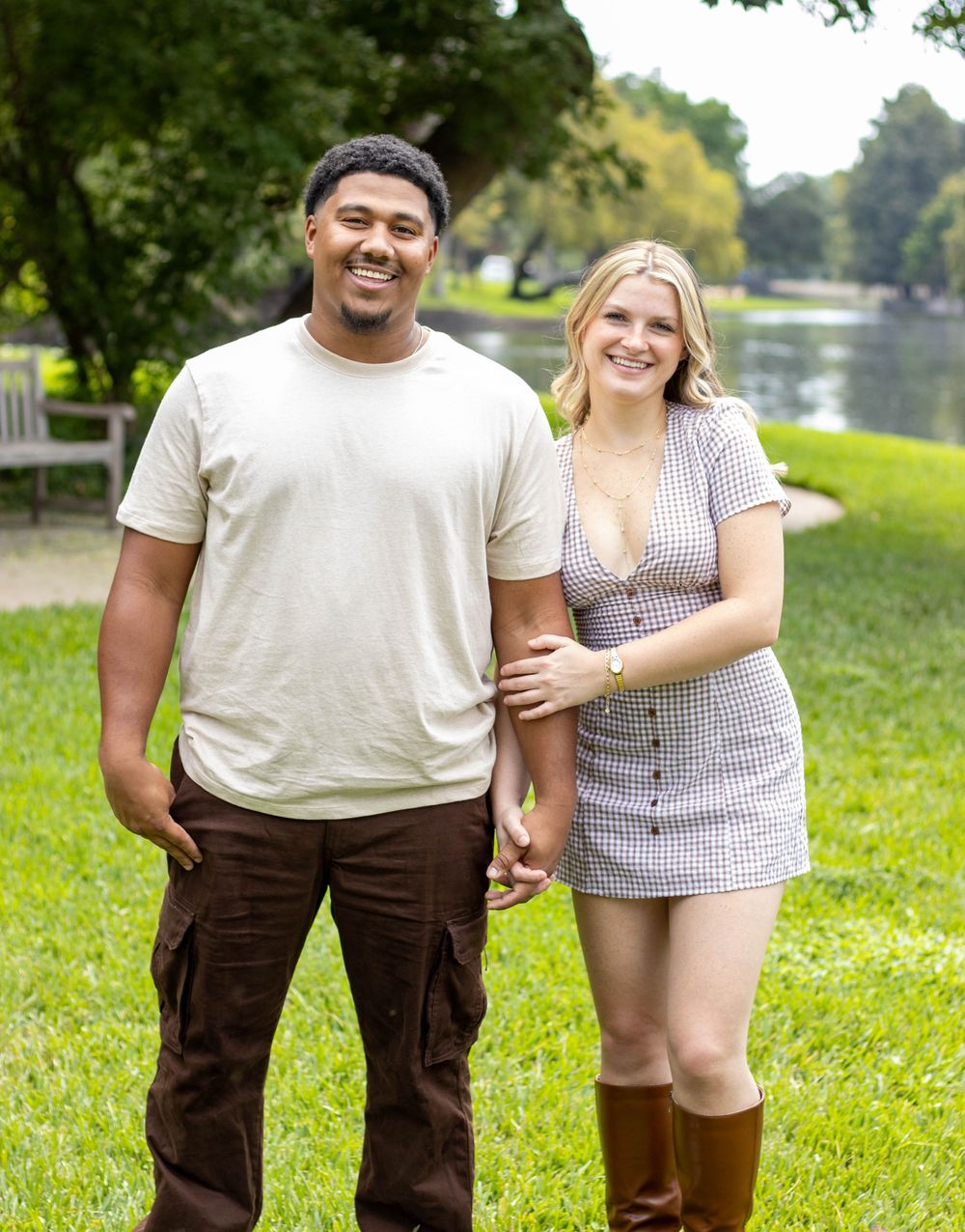 A smiling couple holds hands in a lush green park near a pond, the woman in a plaid mini dress and brown boots.