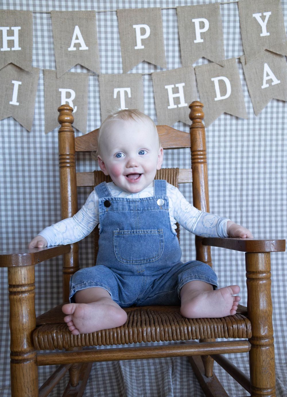 Smiling baby in denim overalls sitting in wooden rocking chair with Happy Birthday burlap banner backdrop.