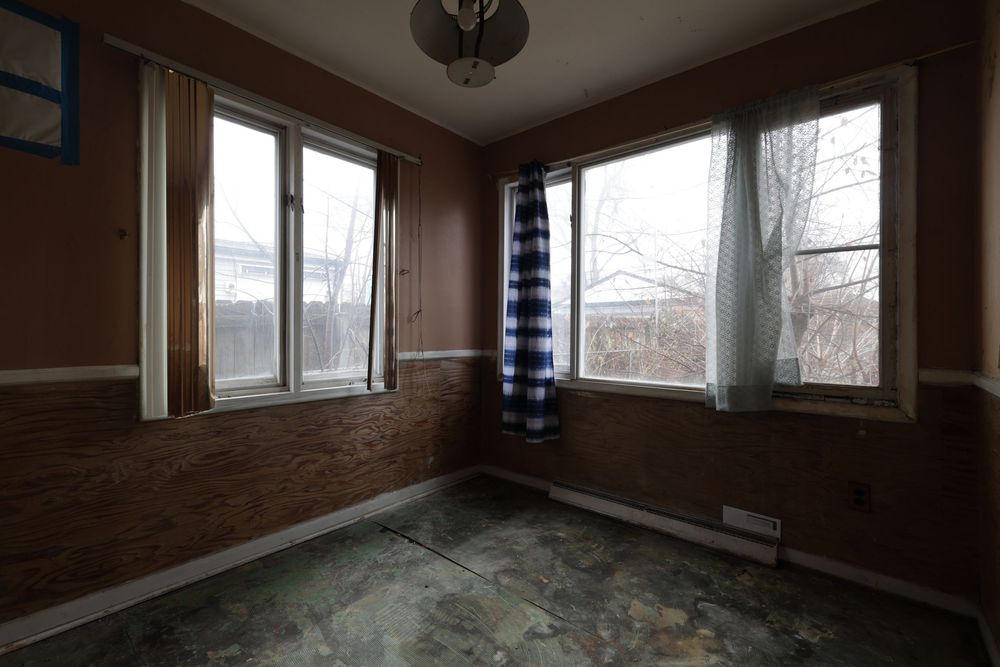 Empty room with brick accent wall, three windows, worn flooring, and a blue plaid curtain in a vacant home.