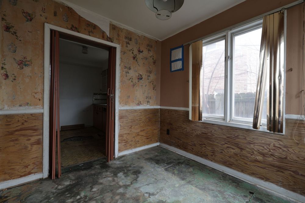 Abandoned room with plywood walls, peeling floral wallpaper, dirty concrete floor, and a window with curtains.