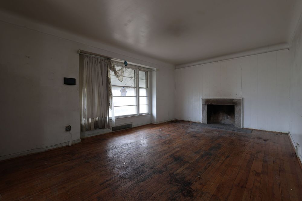 Empty room with hardwood floors, white walls, a fireplace, and a window with sheer curtains in an unfurnished home.