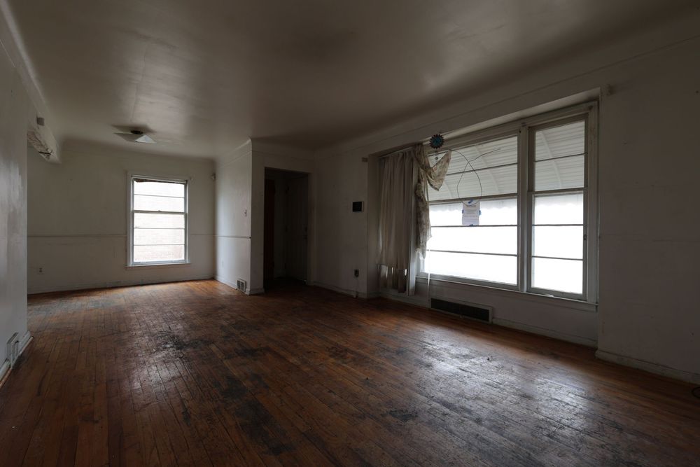 Empty room with hardwood floors, white walls, and two windows allowing natural light in an older home.
